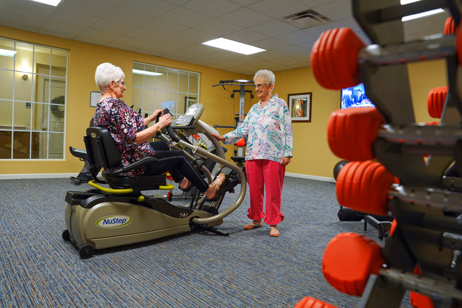 Two elderly women in a fitness room; one is seated on a NuStep exercise machine pedaling, while the other stands nearby watching and smiling. The room has yellow walls, a carpeted floor, a rack of red dumbbells in the foreground, and a television mounted on the wall.