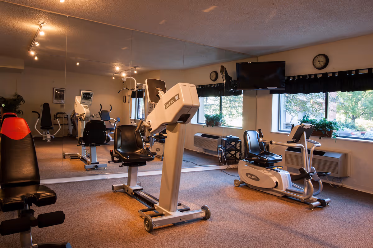 Small fitness room with exercise machines, recumbent bikes, a mirrored wall and windows overlooking trees.