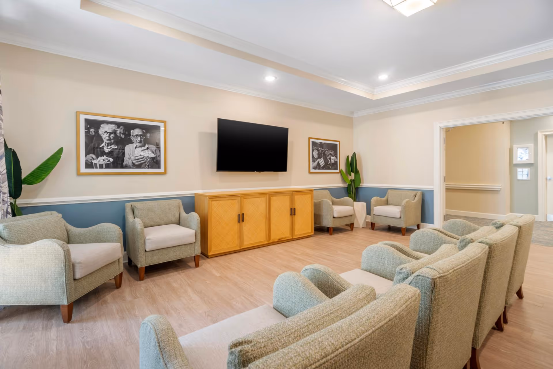 Bright communal seating area with armchairs arranged facing a wall-mounted TV above a wooden cabinet in a memory care living room.
