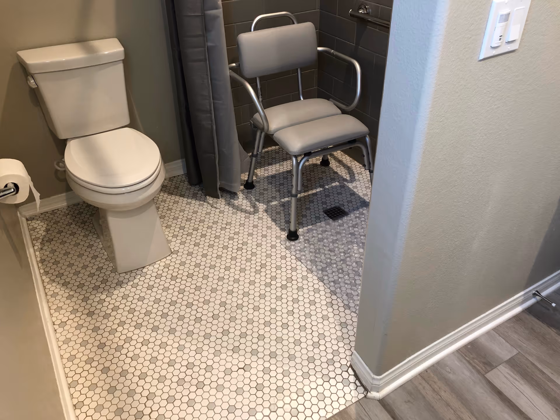 A bathroom with a white toilet, a roll of toilet paper on a holder, and a shower area with a gray shower chair and a gray shower curtain. The floor has small hexagonal tiles in white and gray, and the walls are painted light gray.