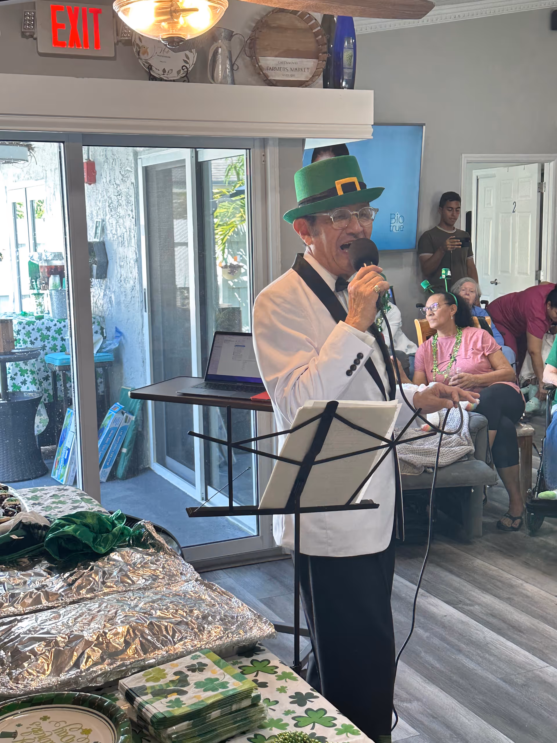 A performer wearing a white jacket and a green leprechaun hat sings into a microphone in a decorated common room while residents watch.