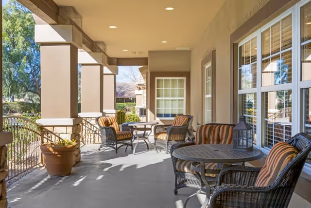 Covered outdoor patio area with several round tables and cushioned wicker chairs arranged for seating. The patio is supported by large columns and overlooks a garden area with trees and shrubs. The building exterior features large windows with white frames and beige walls.