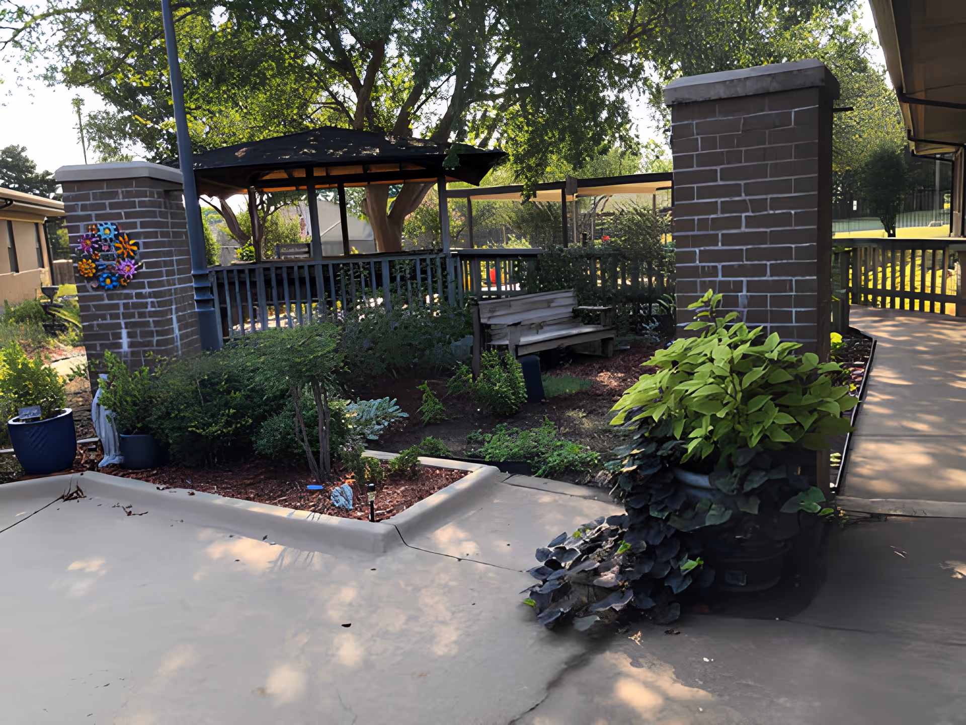Outdoor garden area at a senior living facility with a wooden bench, a gazebo, various plants, and a paved walkway surrounded by greenery and brick pillars.