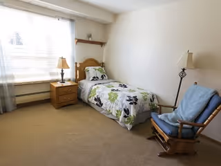 A simple bedroom with a single bed covered in floral bedding, a wooden nightstand with a lamp, a rocking chair with a blue cushion and blanket, a window with sheer curtains, and a small shelf with a plant on the wall.