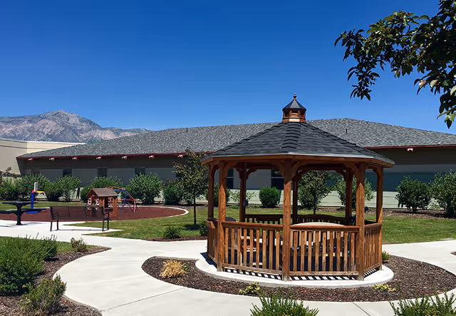 Outdoor area at George E. Wahlen Ogden Veterans Home featuring a wooden gazebo with a shingled roof, surrounded by a paved walkway, green grass, bushes, and trees. In the background, there is a building and mountains under a clear blue sky.