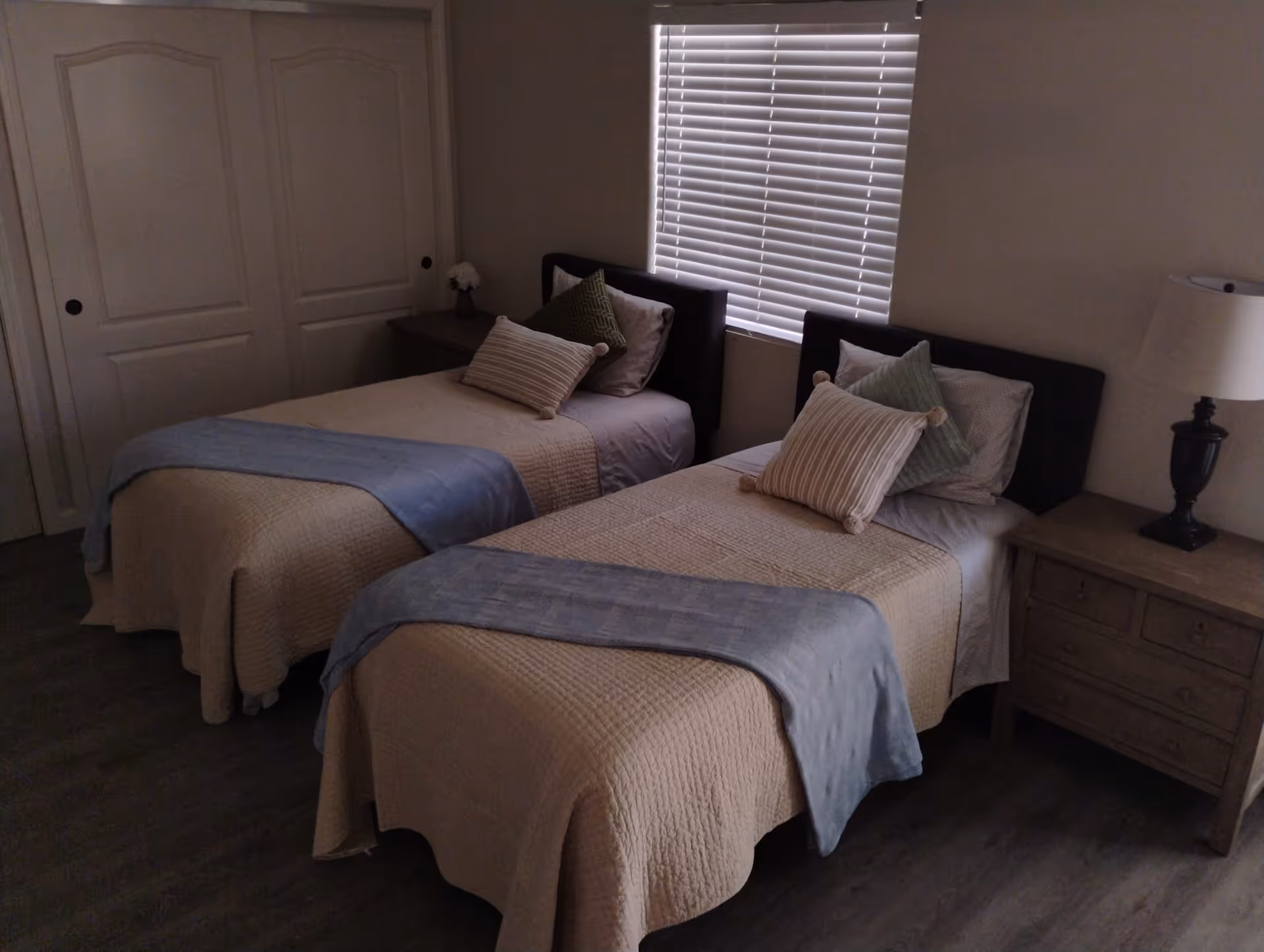 A bedroom with two twin beds, each made with beige quilts and light blue throws at the foot. Each bed has multiple pillows in neutral and green tones. There is a window with closed blinds behind the beds, a nightstand with a lamp on the right side, and a small table with a flower vase on the left side near a closed closet with double doors.