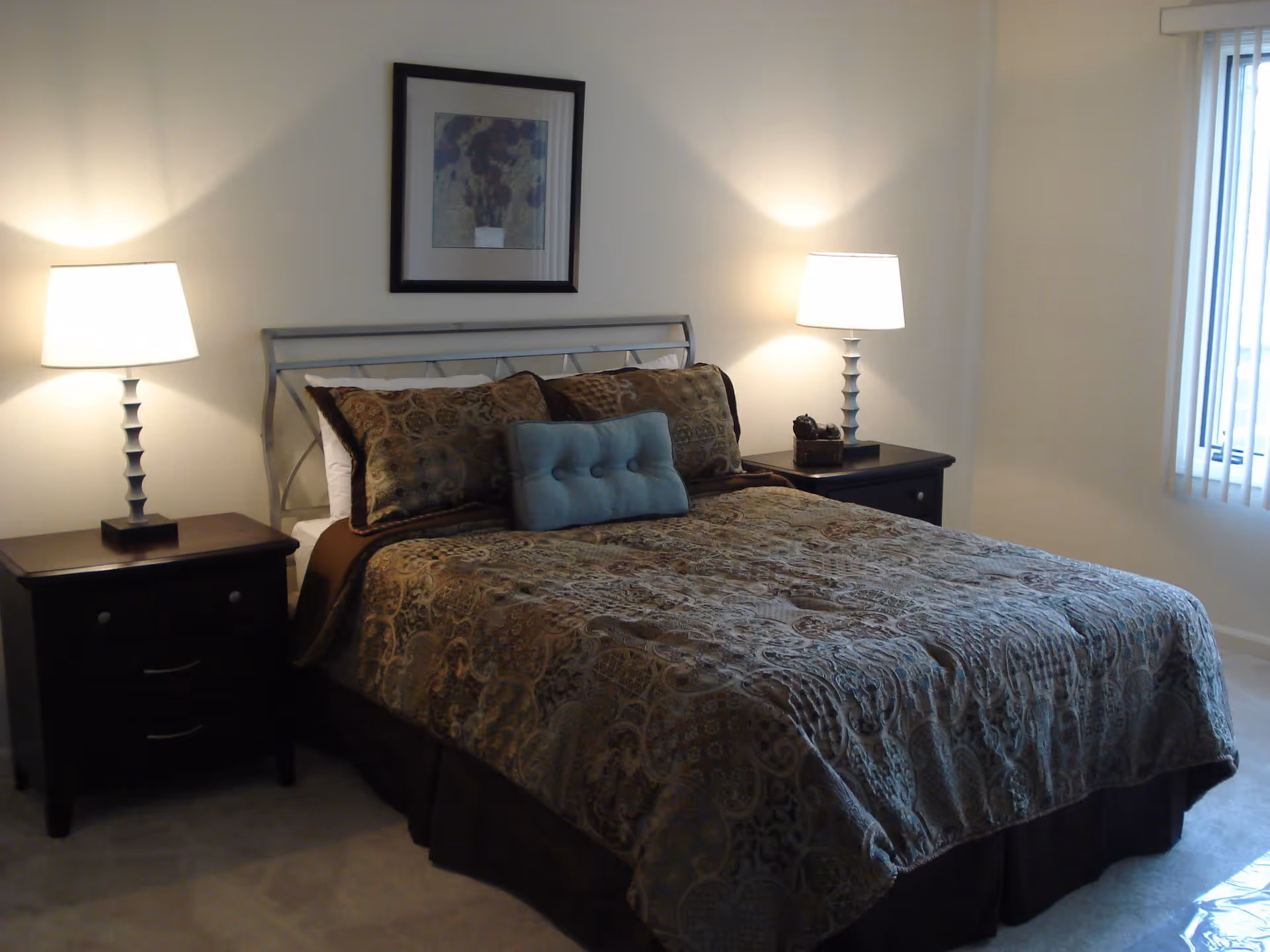 A neatly made bed with patterned brown and beige bedding and a small blue decorative pillow in the center. The bed has a metal headboard and is flanked by two dark wooden nightstands, each with a lit table lamp. A framed picture hangs on the wall above the bed, and there is a window with vertical blinds on the right side of the room.