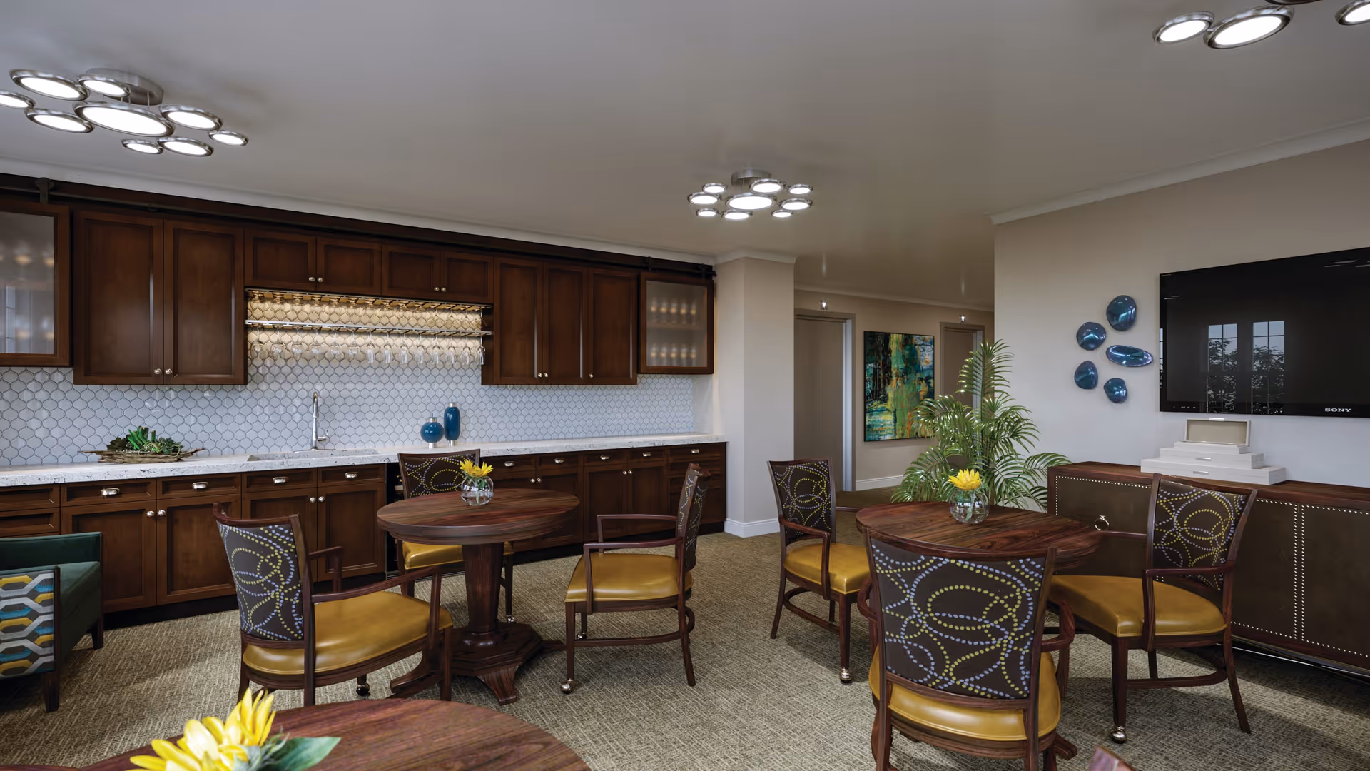 Interior view of a common area with a kitchenette featuring dark wooden cabinets and a white tiled backsplash. There are three round wooden tables with chairs that have patterned backs and yellow seats. A large flat-screen TV is mounted on the wall above a sideboard, and decorative blue wall art is displayed nearby. The room is well-lit with modern ceiling lights and has a carpeted floor.