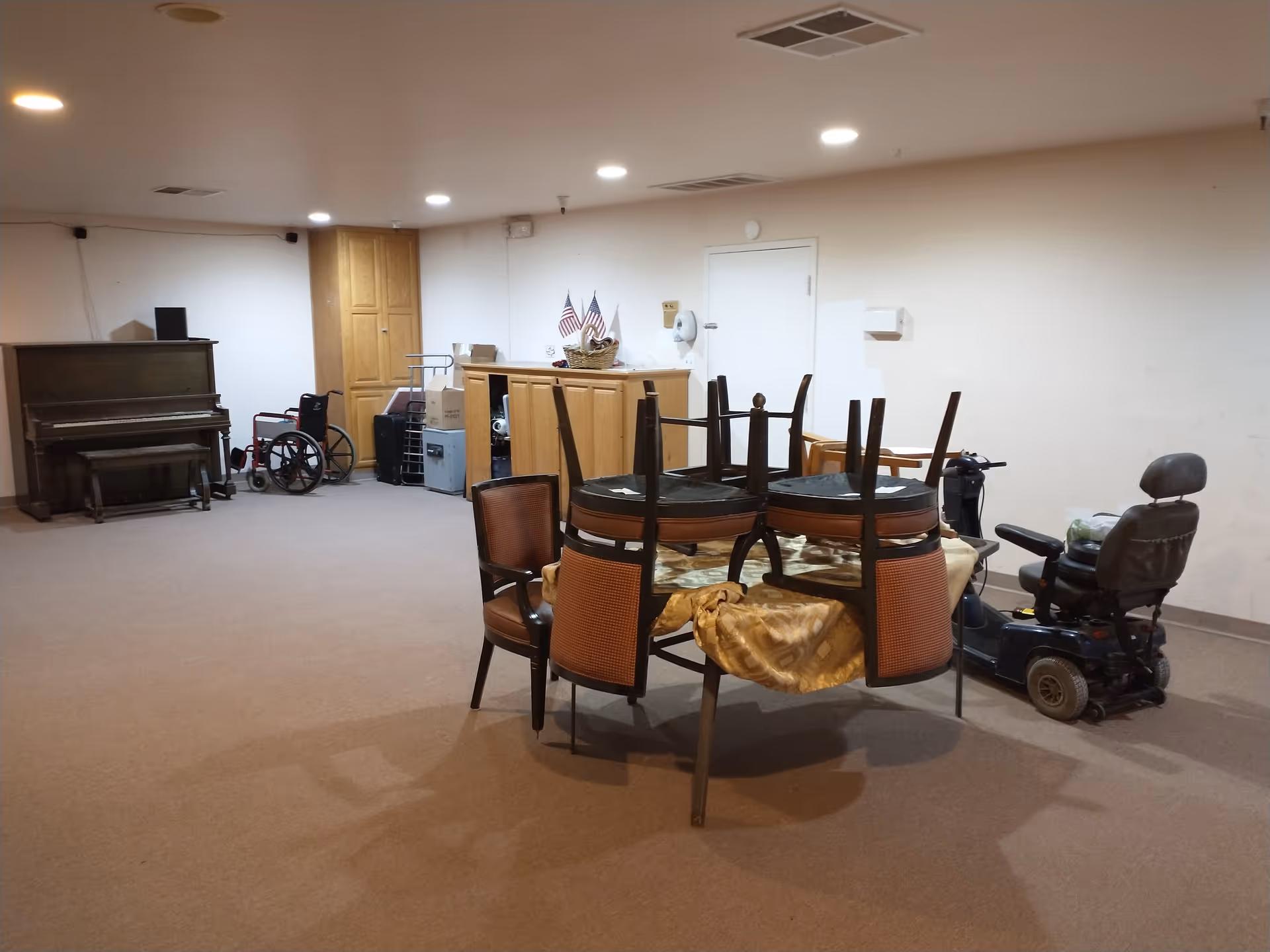 Large interior common room with chairs stacked on a table, a mobility scooter, a piano and storage cabinets.