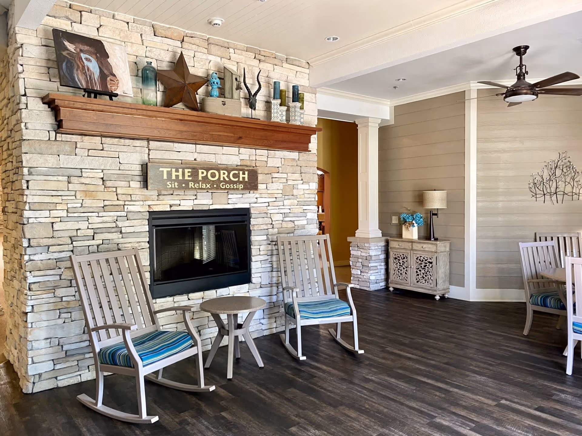 A cozy interior common area with a stone fireplace featuring a wooden mantel decorated with various items including a star, candles, and a painting of a cow. Below the mantel is a sign that reads 'THE PORCH Sit · Relax · Gossip'. In front of the fireplace are two wooden rocking chairs with blue striped cushions and a small round table between them. The room has dark wood flooring, beige walls with white trim, a ceiling fan, and a small cabinet with a lamp and blue flowers on top.