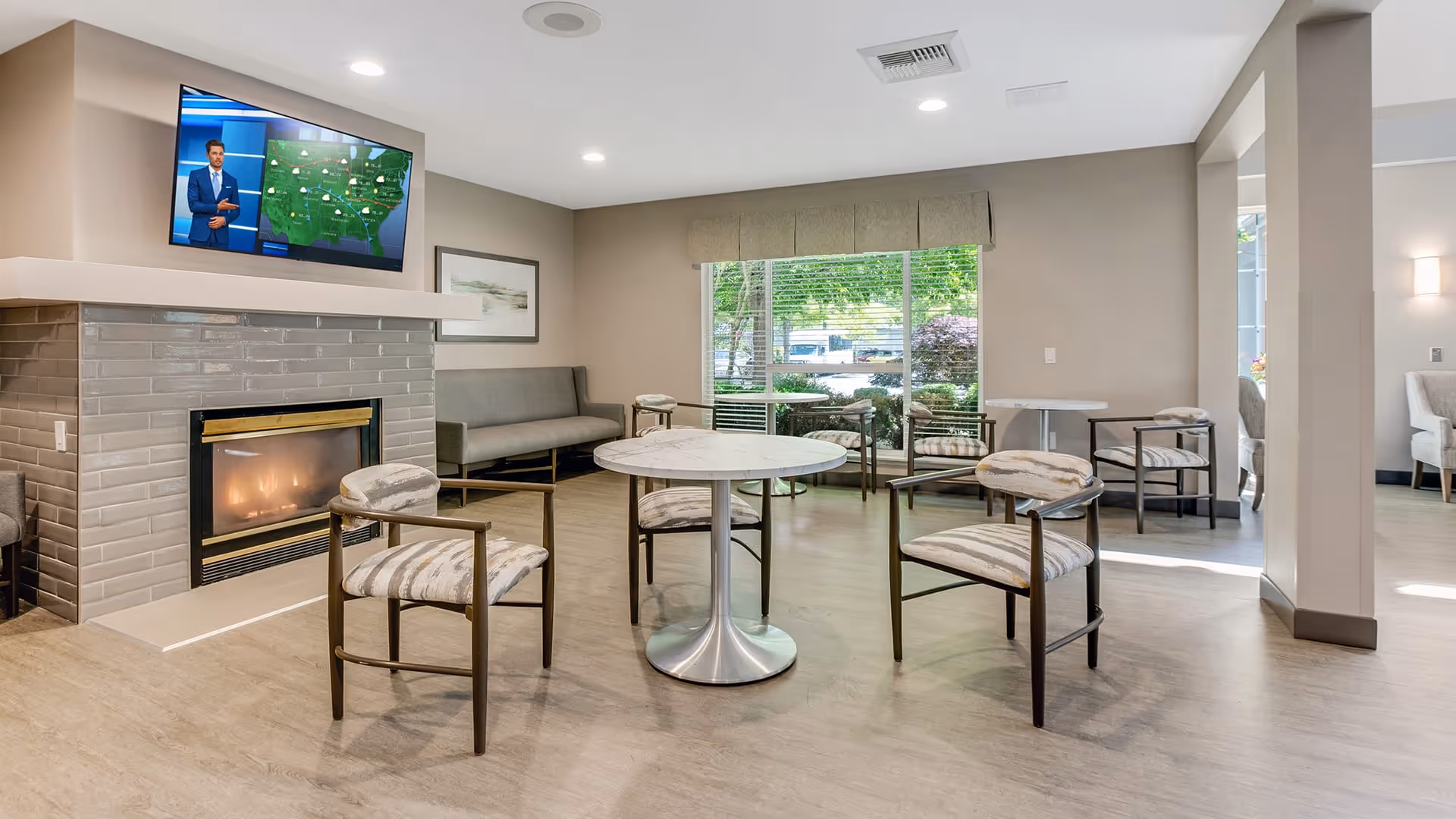 A bright and modern common area with a fireplace, a wall-mounted TV showing a weather forecast, several round tables with cushioned chairs, a long bench along the wall, and large windows letting in natural light with a view of greenery outside.