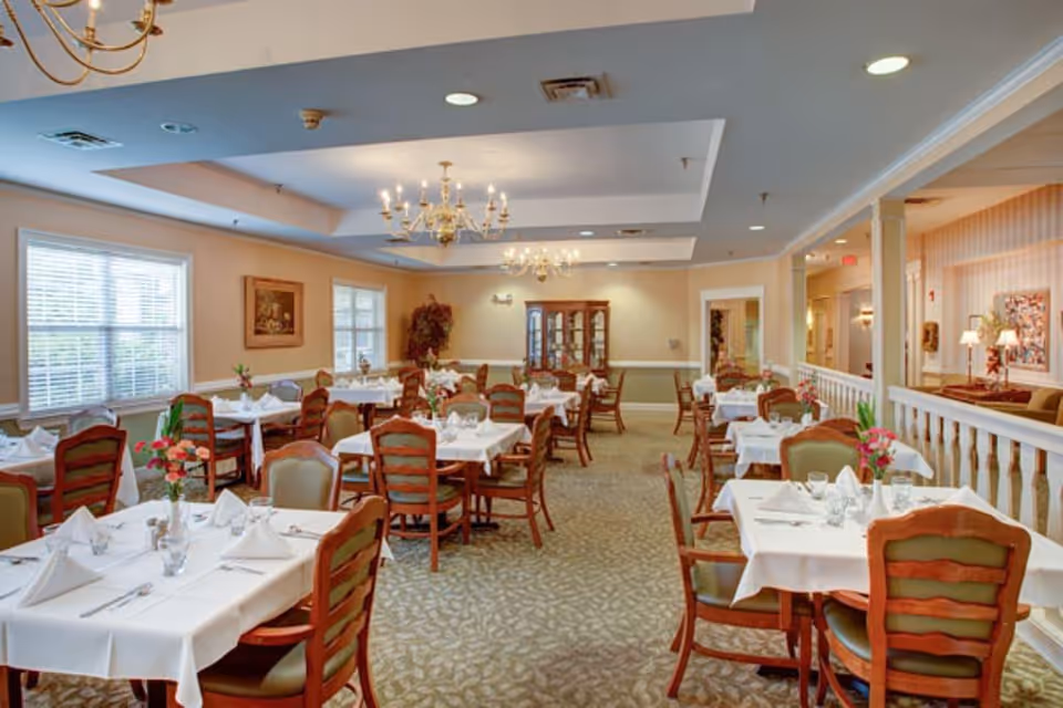 A well-lit dining room with multiple tables covered in white tablecloths, each set with napkins, glasses, and silverware. Wooden chairs with green cushions surround the tables. The room features beige walls, large windows with blinds, chandeliers hanging from the ceiling, and a china cabinet against the far wall. There are floral centerpieces on each table and a carpeted floor with a subtle pattern.