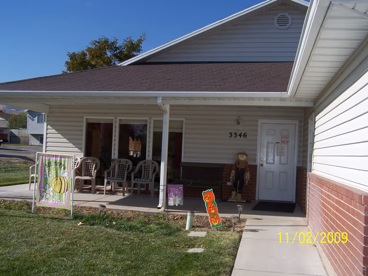 Front porch of a residential building with the address number 3346. The porch has several plastic chairs, a bench, and seasonal decorations including a scarecrow and signs with a bee and pumpkins. The building has beige siding with a brick lower section and a white door. There is a grassy lawn in front and a concrete walkway leading to the porch.