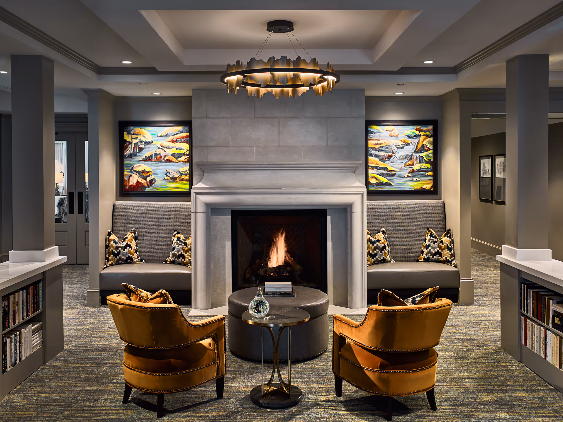 A cozy living room area featuring a lit fireplace with a stone mantel. Two mustard yellow armchairs face the fireplace, separated by a small round table with a decorative glass object. Behind the fireplace are two built-in gray cushioned benches with patterned pillows. Colorful landscape paintings hang above each bench. The room has a modern chandelier and built-in bookshelves on either side of the seating area.