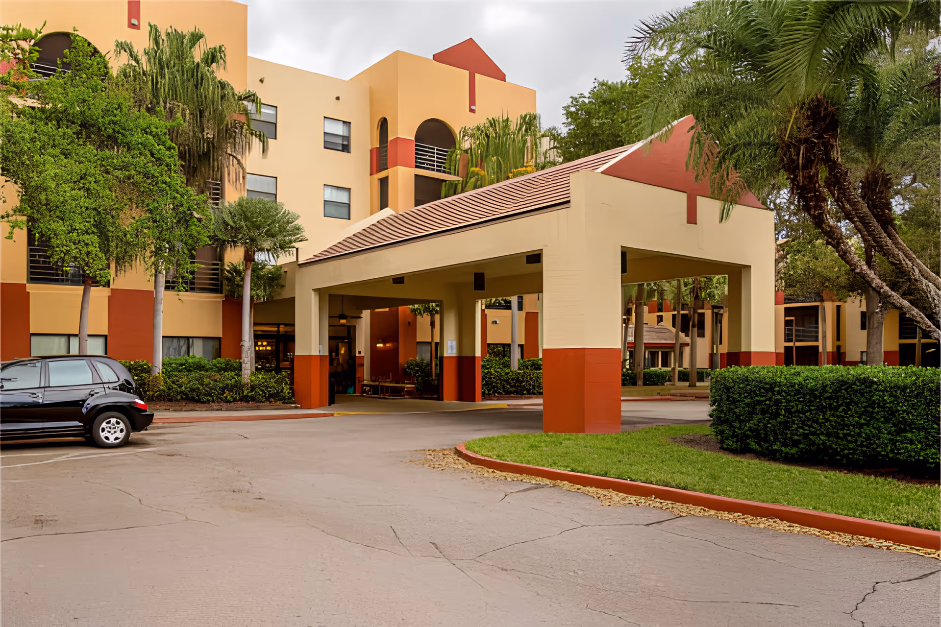Exterior view of Elison Assisted Living of Bella Vita showing the front entrance with a covered drop-off area, surrounded by palm trees and greenery. The building is painted in beige and red tones with multiple windows and balconies.