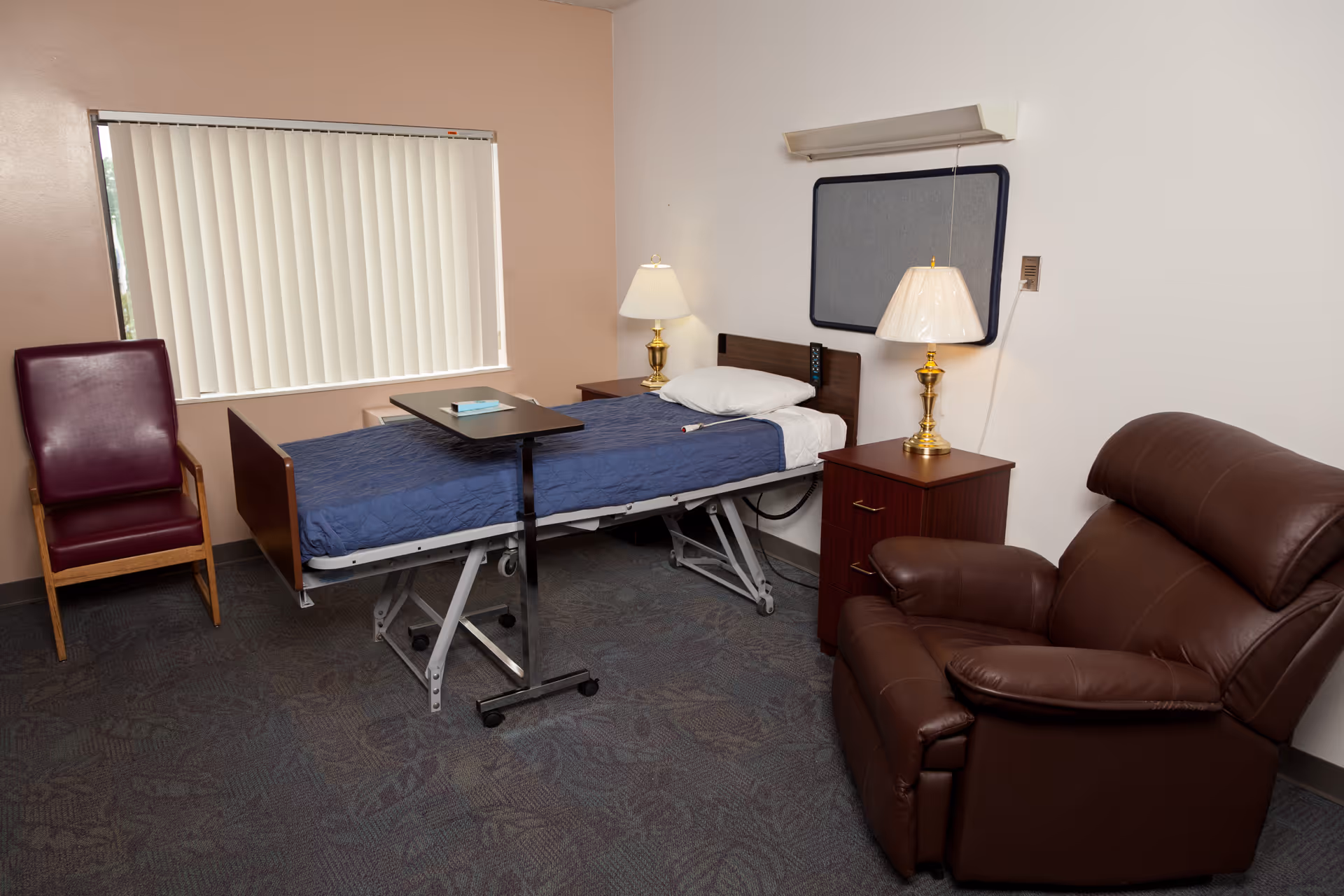A patient room in a healthcare facility featuring a hospital bed with a blue blanket, an overbed table with a book on it, a wooden nightstand with a brass lamp, a brown leather recliner chair, a maroon chair, and a window with vertical blinds.