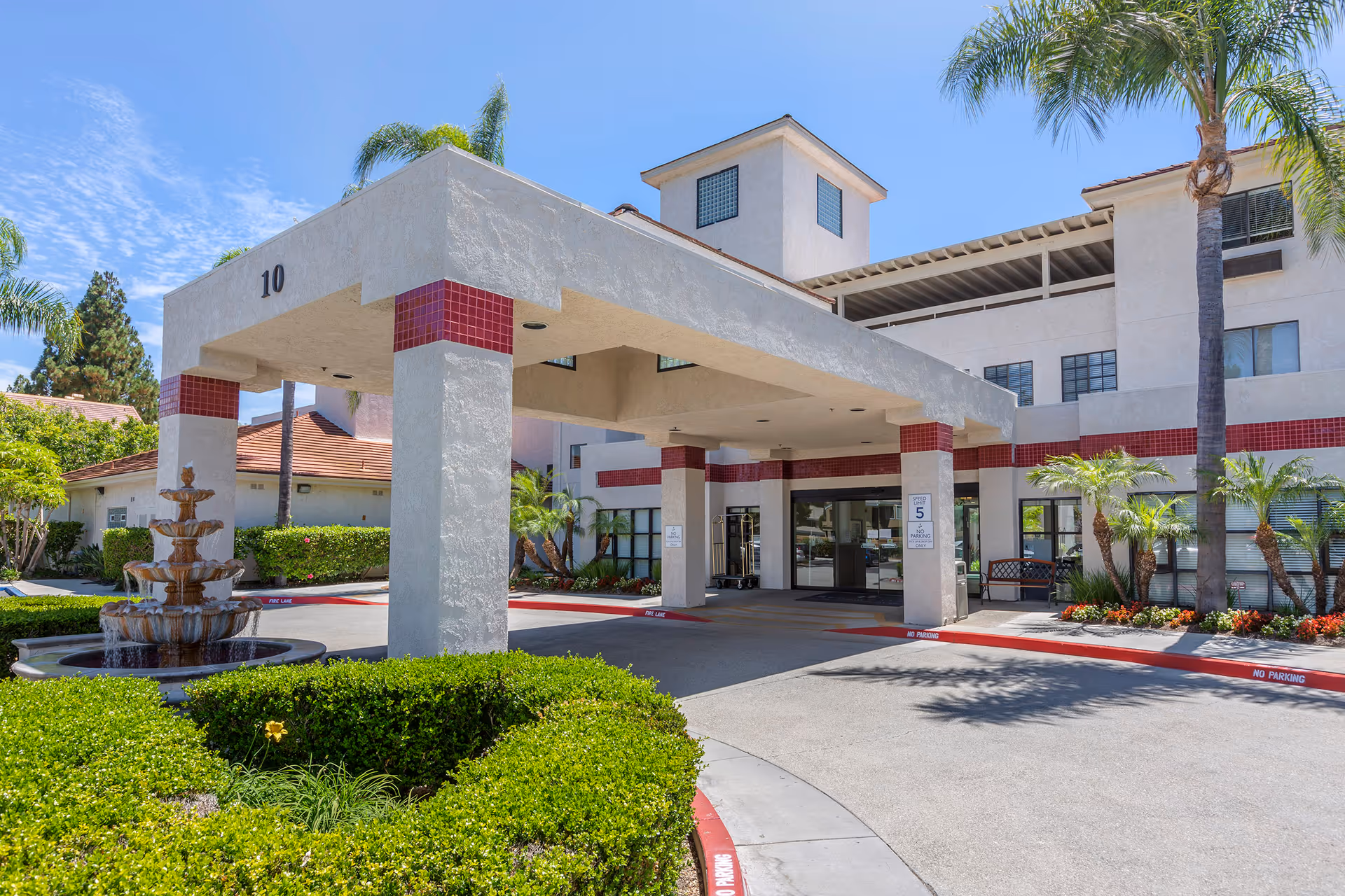 Exterior view of Brookdale Irvine senior living facility entrance with a covered drop-off area supported by columns with red tile accents, a tiered water fountain surrounded by greenery, palm trees, and clear blue sky.