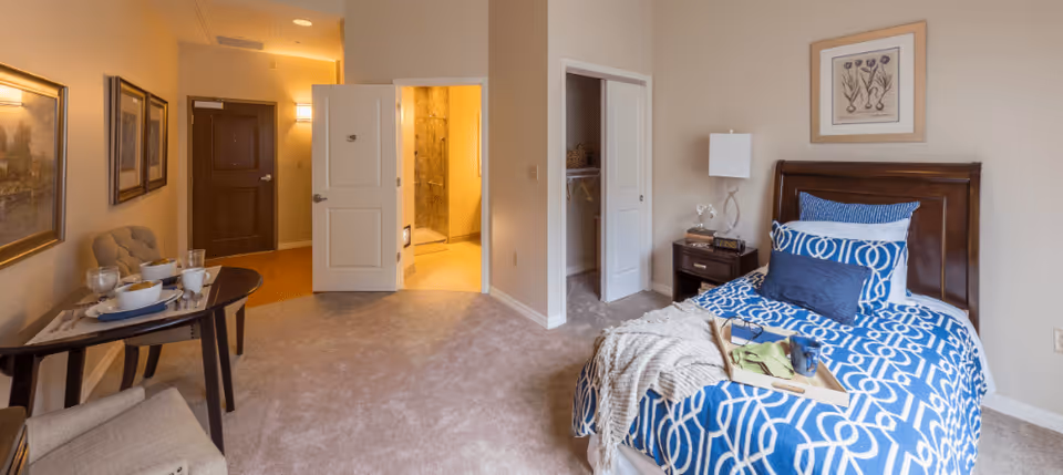 A cozy bedroom in a senior living facility with a single bed dressed in blue and white patterned bedding, a wooden nightstand with a lamp and clock, an open closet, a small dining table set with dishes and cups, and a view into a bathroom with a shower.