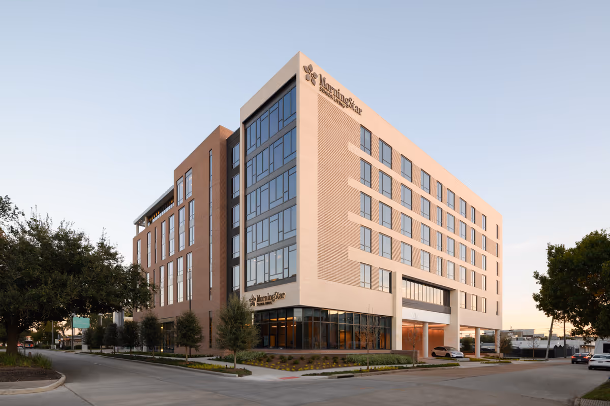 Exterior view of a modern multi-story building with large windows and a mix of brick and light-colored facade. The building is identified as MorningStar Senior Living at River Oaks, with the name visible on the upper corner and near the entrance. There are trees and landscaping around the building, and a few cars are parked nearby under a clear sky.