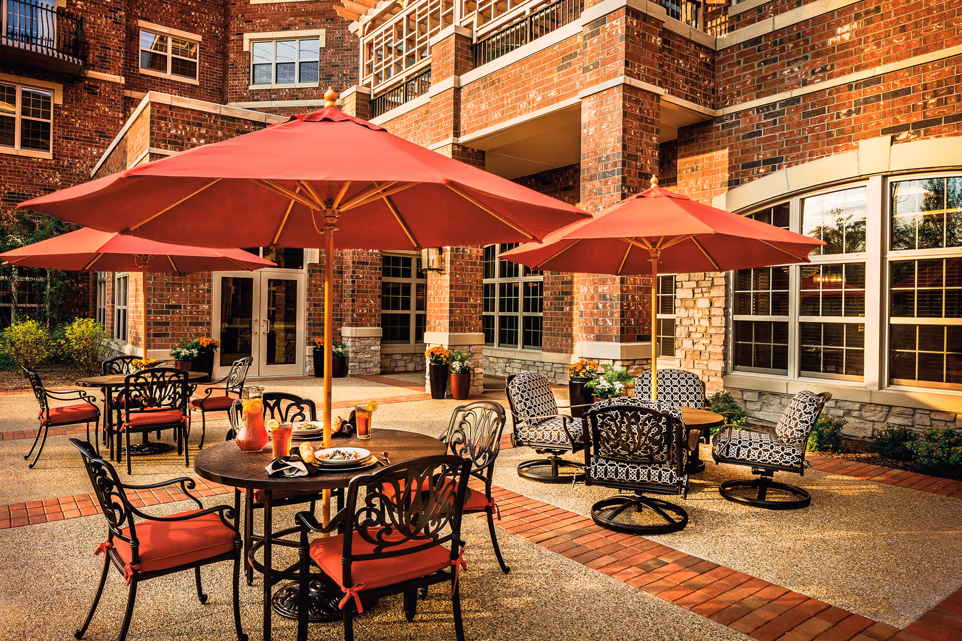 Outdoor patio area at North Shore Place with round tables, red umbrellas, and chairs with red cushions and patterned upholstery. The patio is surrounded by a brick building with large windows and potted plants.