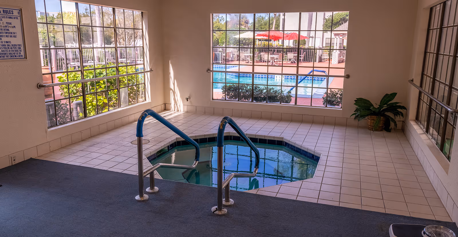 Indoor hot tub with tiled floor and metal handrails, large windows showing an outdoor swimming pool area with tables, chairs, and red umbrellas, and a potted plant in the corner.