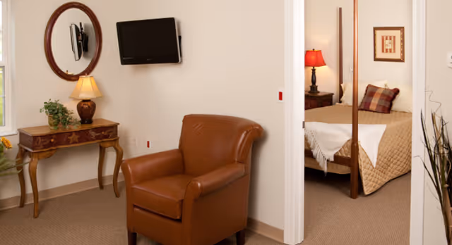 A cozy interior space featuring a brown leather armchair in the foreground, a small wooden table with a lamp and a plant against the wall, a round mirror above the table, and a wall-mounted flat screen TV. Through an open doorway, a bedroom is visible with a bed covered in a beige quilt, a red lamp on a nightstand, and a framed picture on the wall.