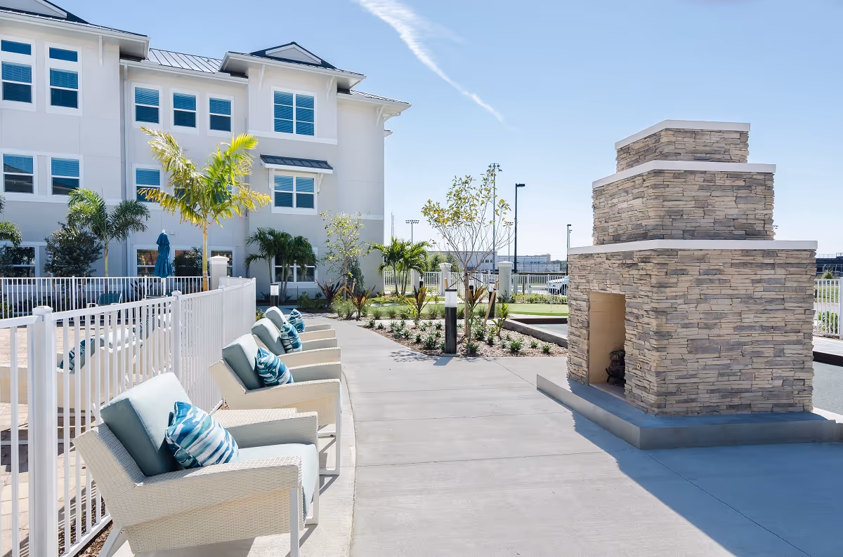 Outdoor seating area at American House Fort Myers featuring cushioned wicker chairs with blue and white pillows, a stone outdoor fireplace, palm trees, and a white fence surrounding the space under a clear blue sky.
