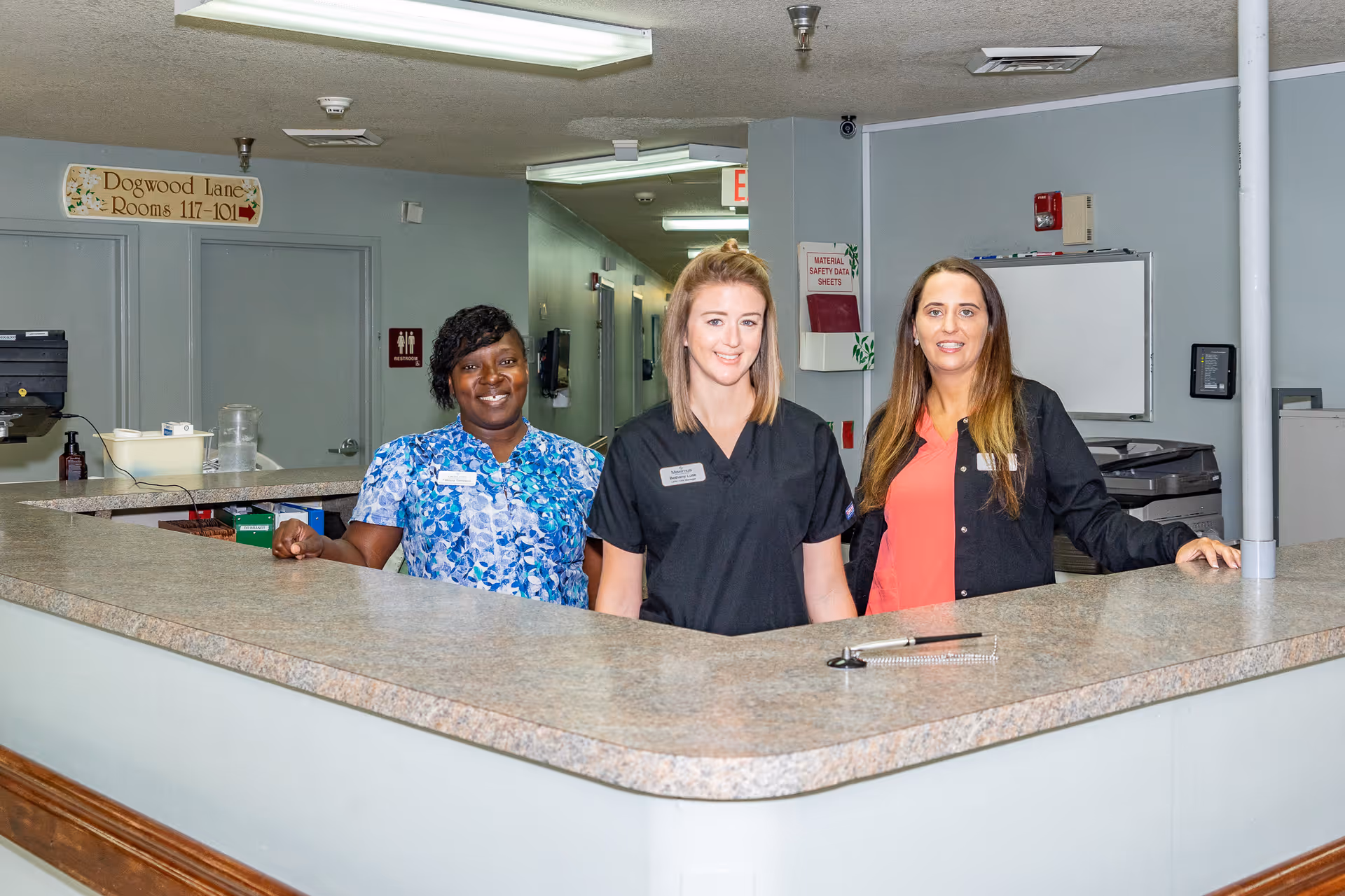 Three female staff members standing behind a reception desk in a nursing home facility. The background shows a hallway with a sign indicating rooms 117-101 and a restroom sign.