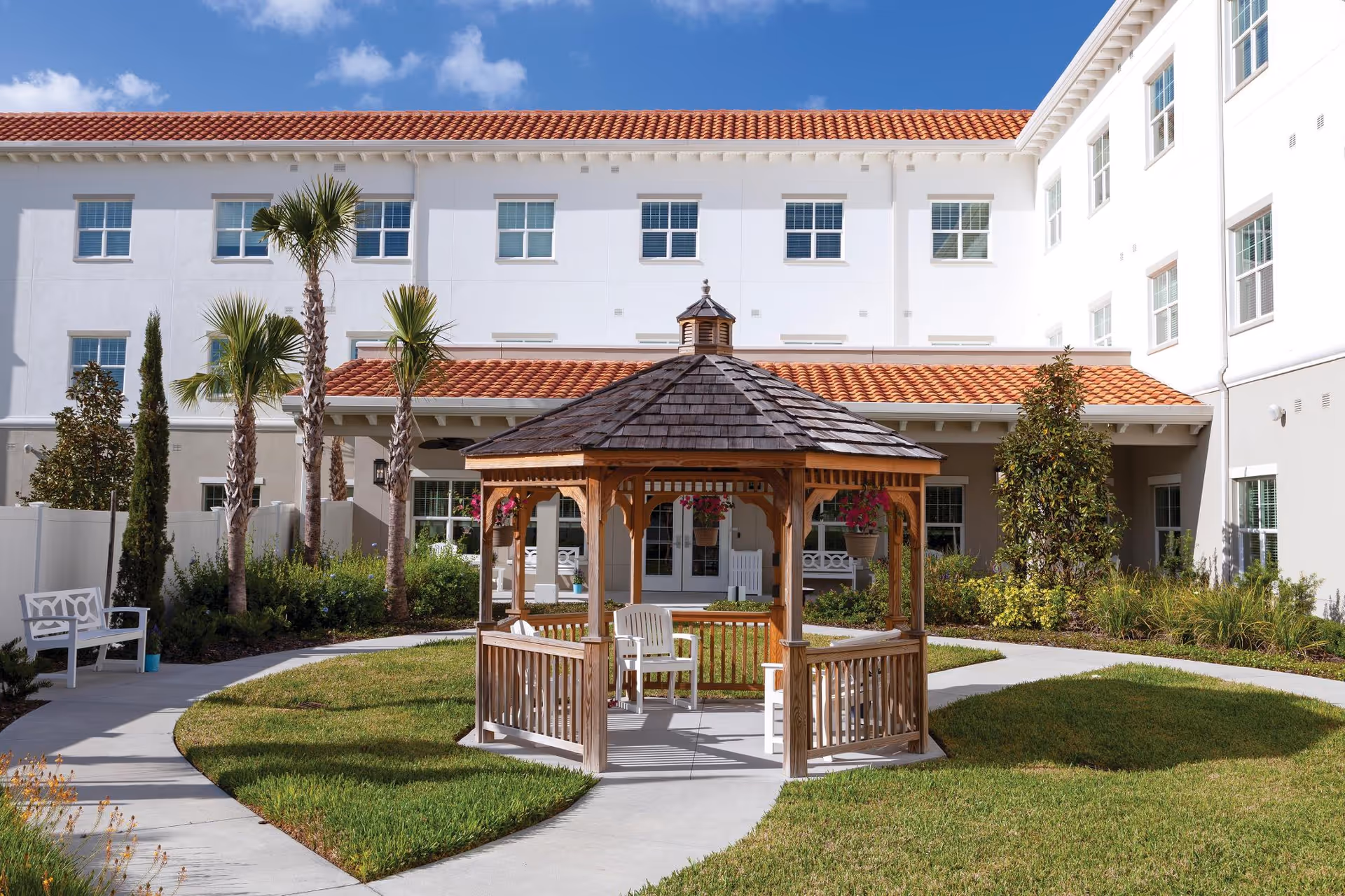 A landscaped courtyard with a wooden gazebo, walkways, benches, and a white multi-story building in the background.