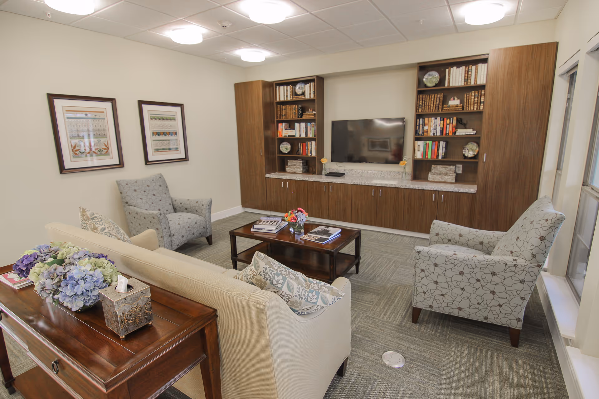 A cozy living room area with a beige sofa adorned with patterned pillows, two patterned armchairs, a wooden coffee table with books and a small flower arrangement, and a wooden console table with a floral centerpiece and a decorative box. The back wall features built-in wooden shelves filled with books and decorative items, and a flat-screen TV mounted in the center. Two framed artworks hang on the adjacent wall, and a window allows natural light into the room.