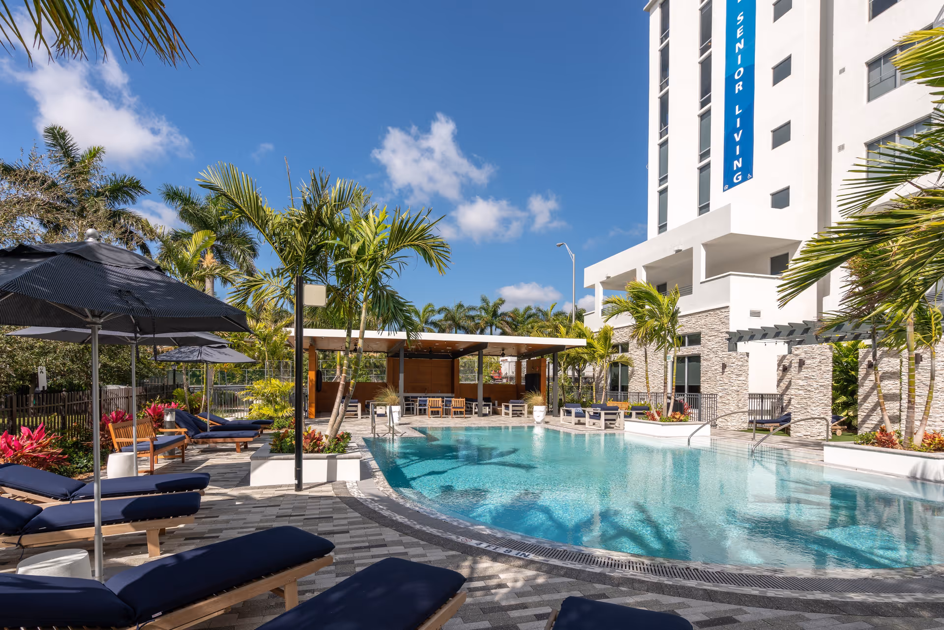 Outdoor swimming pool area at a senior living facility with lounge chairs, umbrellas, palm trees, and a covered seating area under a clear blue sky.