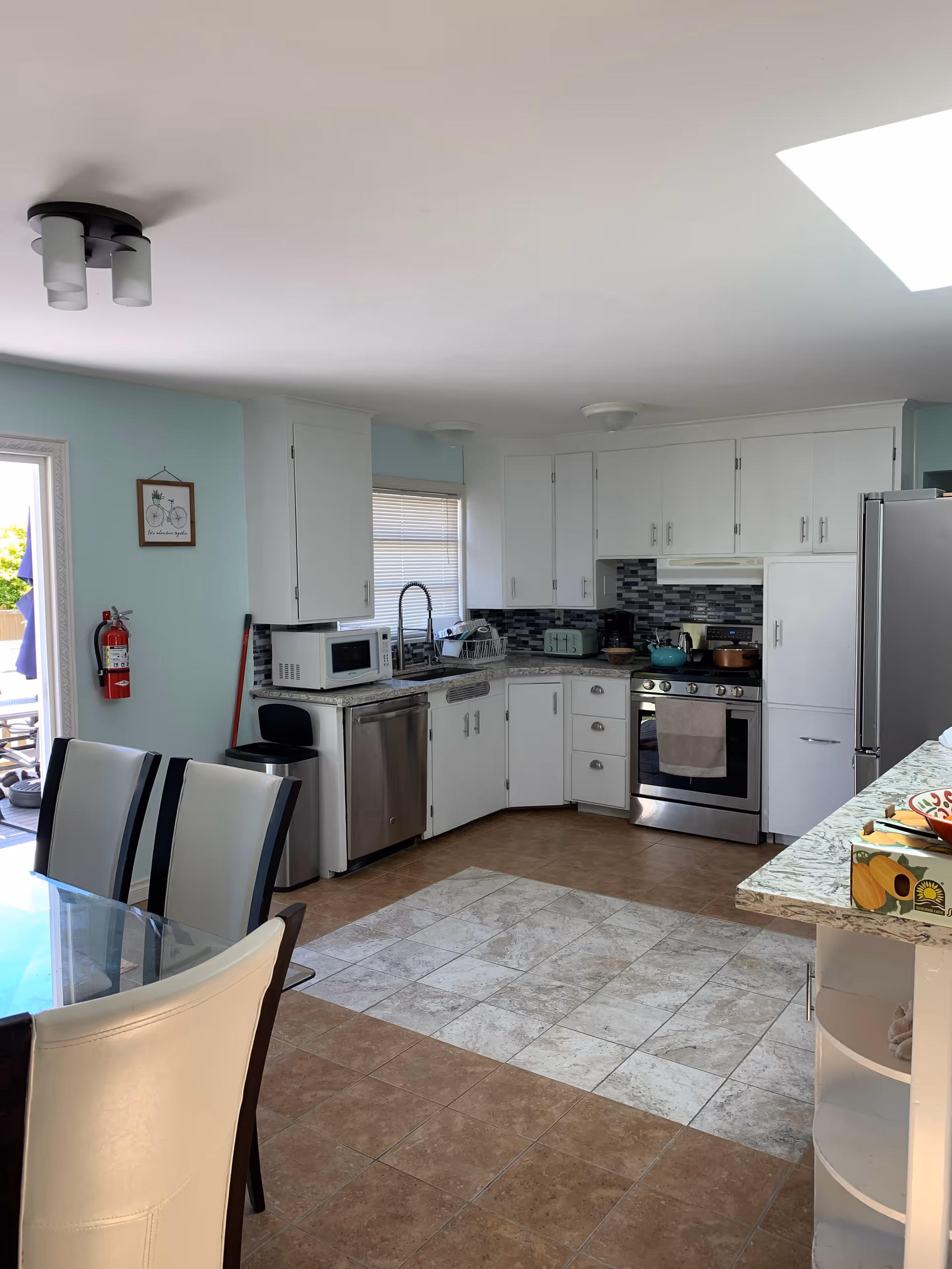 A bright kitchen with white cabinets, stainless steel appliances including a dishwasher, stove, and refrigerator. There is a glass dining table with white and black chairs in the foreground. The kitchen has a tiled floor with a lighter tile section in the center, a window with blinds above the sink, and a fire extinguisher mounted on a light blue wall near the door leading outside.