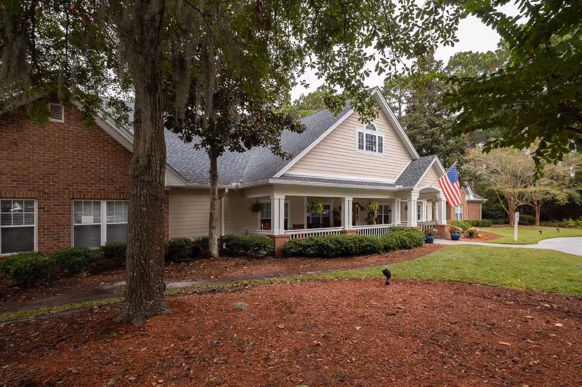 Exterior view of a single-story residential-style building with beige siding and brick accents, a covered porch with white railings, hanging plants, and an American flag. The building is surrounded by trees, shrubs, and a mulched garden area with a lawn and a curved driveway.