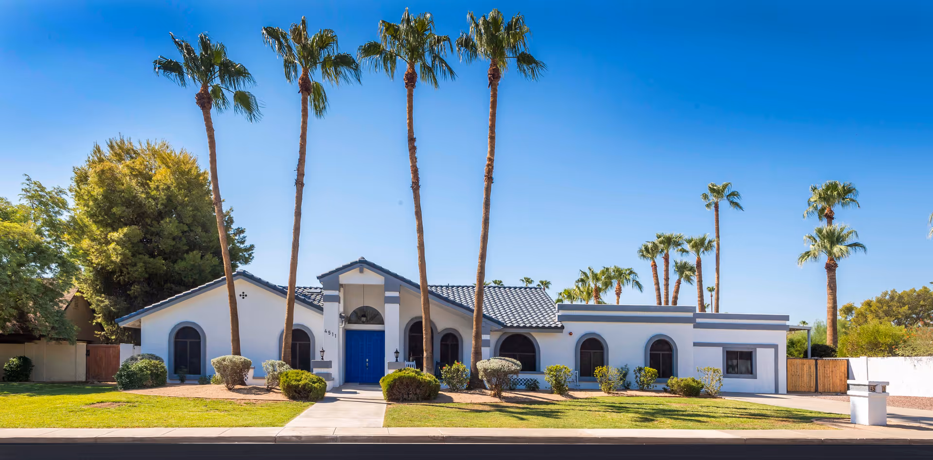 Front exterior view of Bella Vita Assisted Living at Cholla, a single-story building with a blue door, arched windows, and several tall palm trees in the front yard under a clear blue sky.