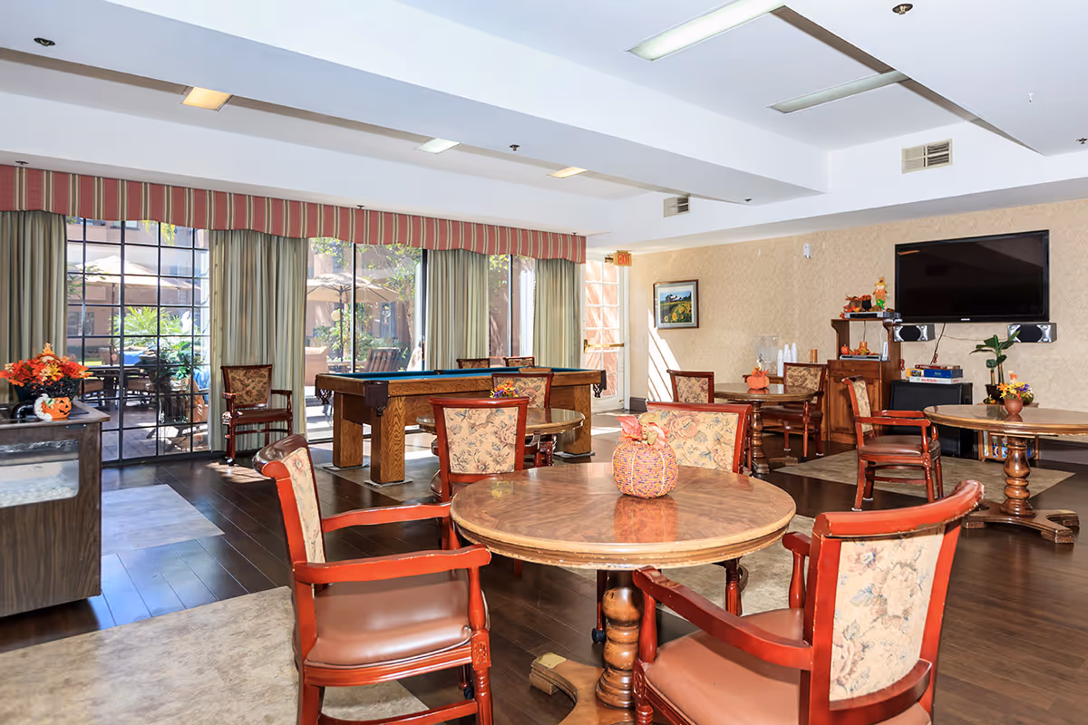 A bright and spacious common area in a senior living facility with round wooden tables and upholstered chairs arranged around the room. A pool table is positioned near large windows with curtains, allowing natural light to fill the space. The room features a wall-mounted flat-screen TV, a small cabinet with books and decorations, and a door leading to an outdoor patio area with tables and umbrellas.