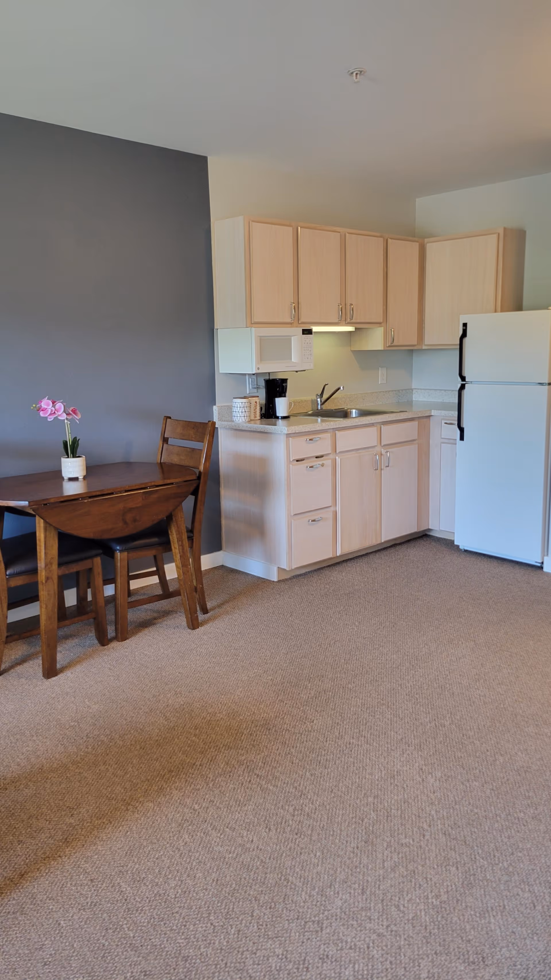 A small kitchen area with light wood cabinets, a white refrigerator, a microwave, a coffee maker, and a sink. Next to the kitchen is a wooden dining table with two chairs and a small potted orchid on the table. The wall behind the table is painted gray, and the floor is carpeted.