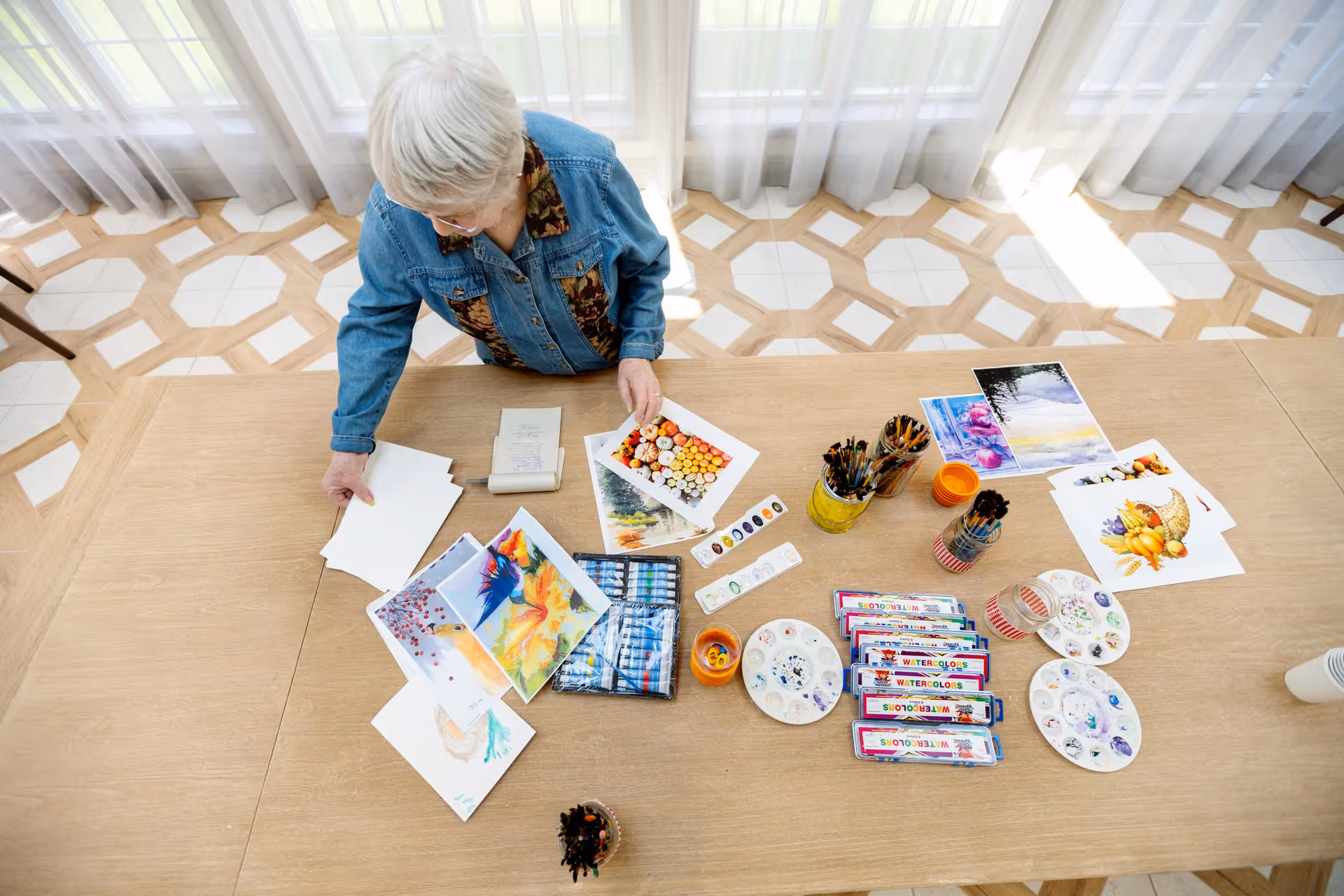 An elderly woman with short white hair wearing a denim jacket is standing at a wooden table covered with watercolor paintings, paint palettes, brushes in jars, and watercolor paint sets. The room has large windows with sheer white curtains and a patterned floor.