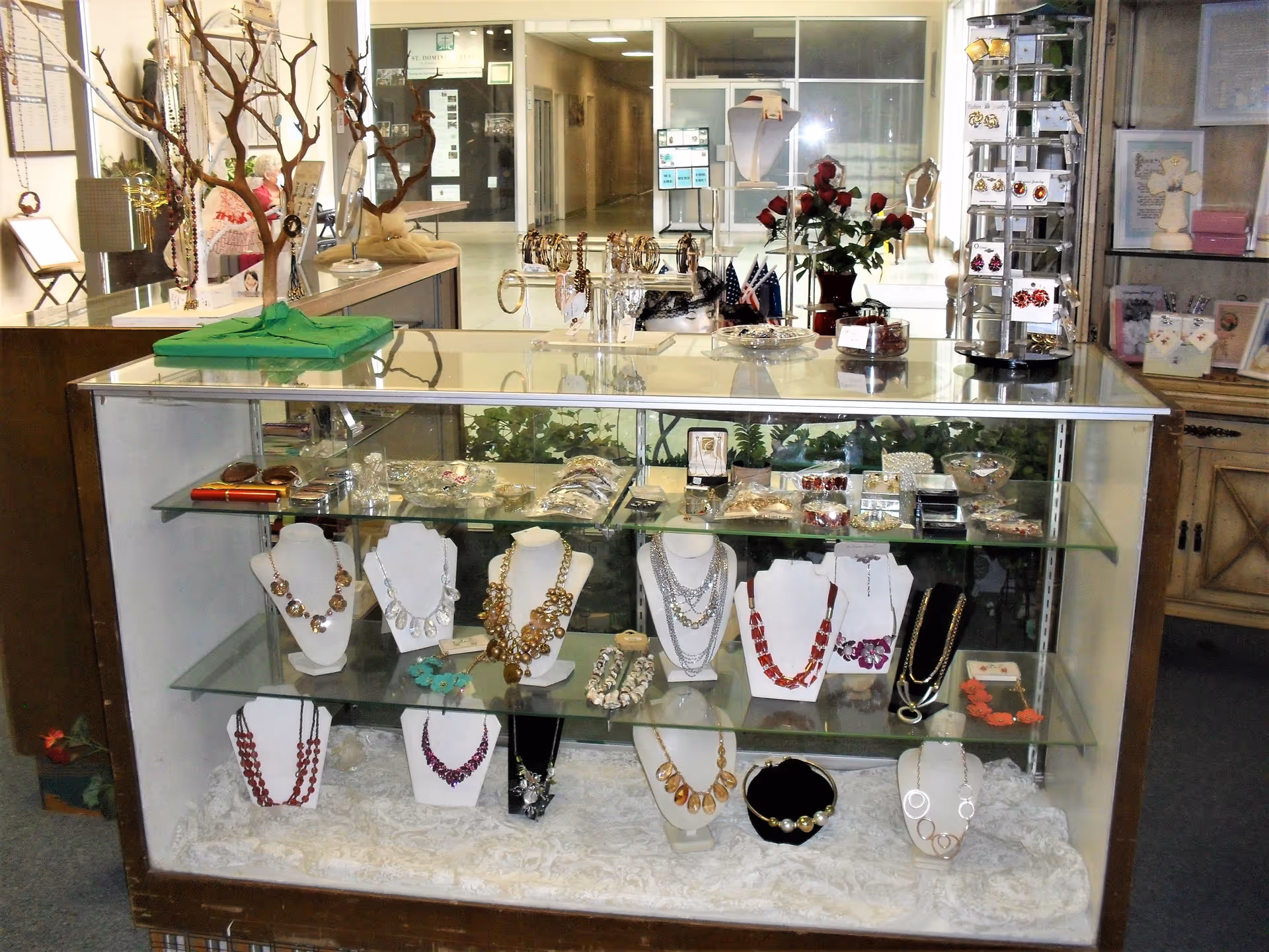 Glass display case filled with various necklaces, bracelets, and jewelry pieces inside a retail area of a senior living facility. The background shows a hallway and additional display items including a vase with red roses and a rotating jewelry stand.