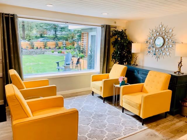 A cozy living room with four yellow armchairs arranged around a small round table with a flower vase. The room features a large window overlooking a green garden with outdoor chairs. There is a decorative mirror on the wall above a dark blue sideboard with two lamps and a potted plant in the corner.