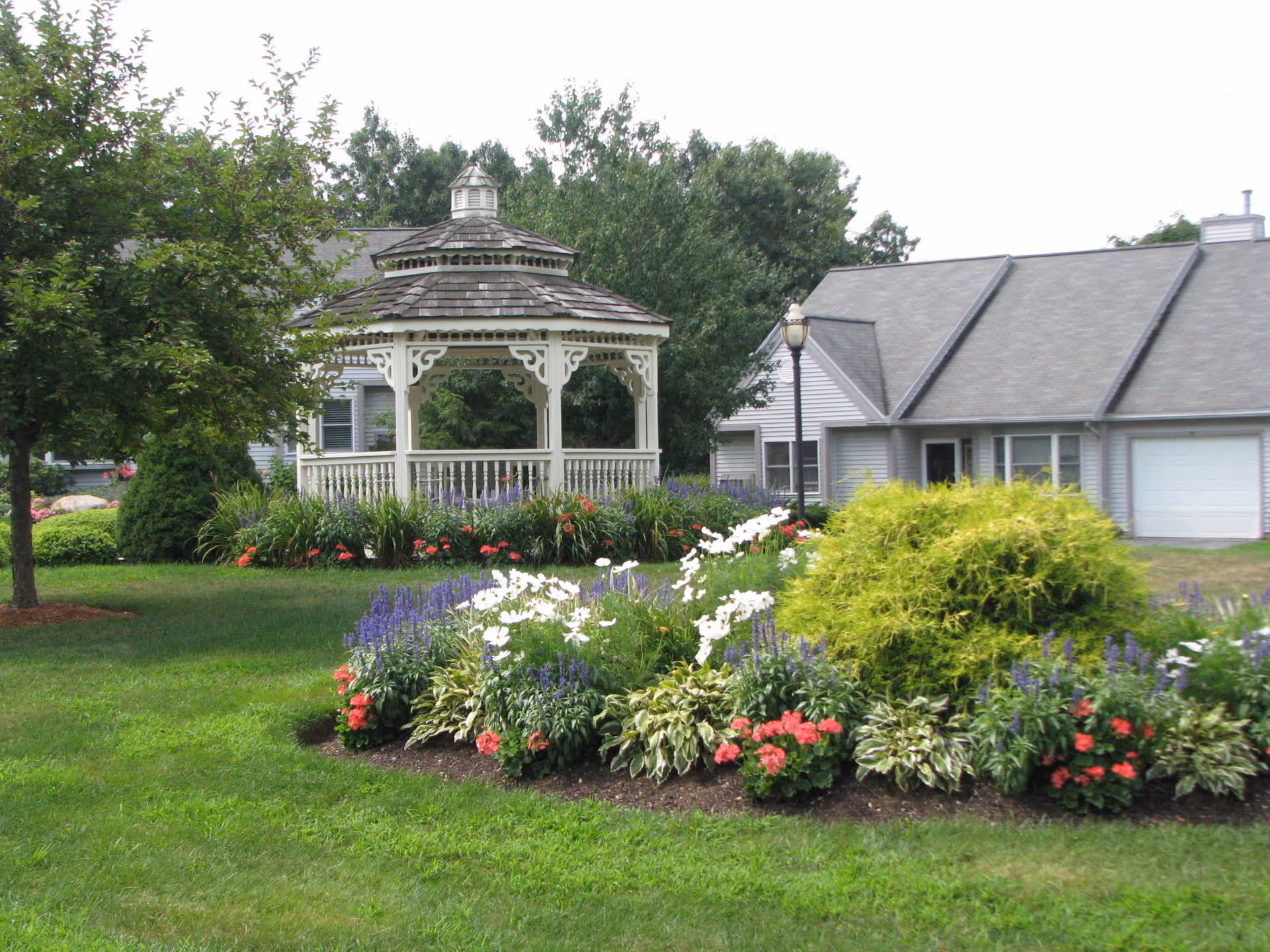 A well-maintained garden area with colorful flowers including white, purple, and red blooms, a green bush, and a tree on the left. In the background, there is a white wooden gazebo with decorative trim and a shingled roof. Behind the gazebo, there is a gray building with a sloped roof and a garage door.