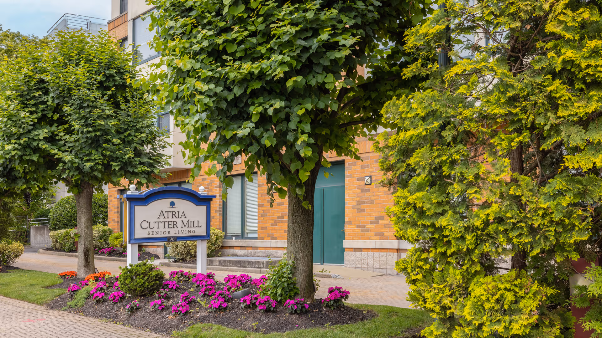Entrance of Atria Cutter Mill senior living building with a sign, trees, and a colorful flowerbed.