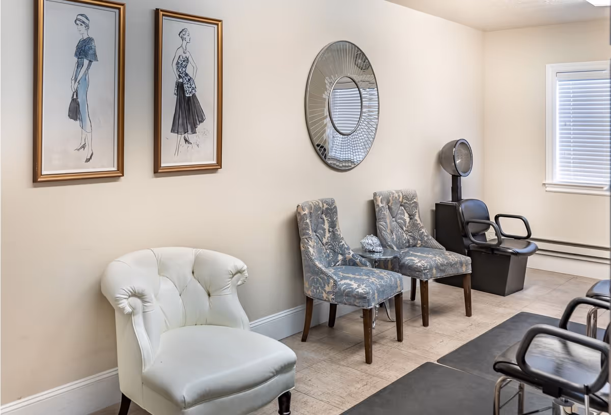 A waiting area with a white cushioned chair, two patterned blue chairs, a small round table, a round decorative mirror on the wall, two framed fashion sketches, and a vintage hair dryer chair near a window with blinds.