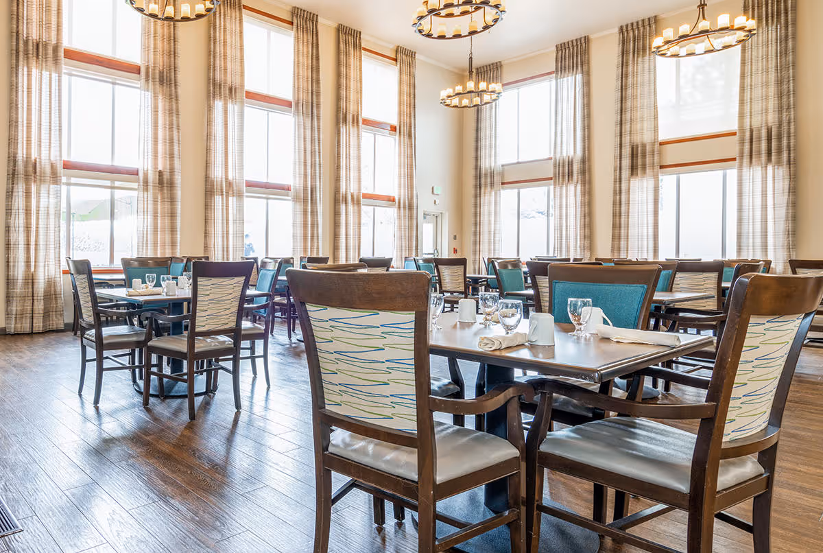 A bright dining room with multiple tables and chairs arranged neatly. The room features large windows with tall plaid curtains allowing natural light to fill the space. Tables are set with glasses, napkins, and cups, and there are two circular chandeliers hanging from the ceiling.