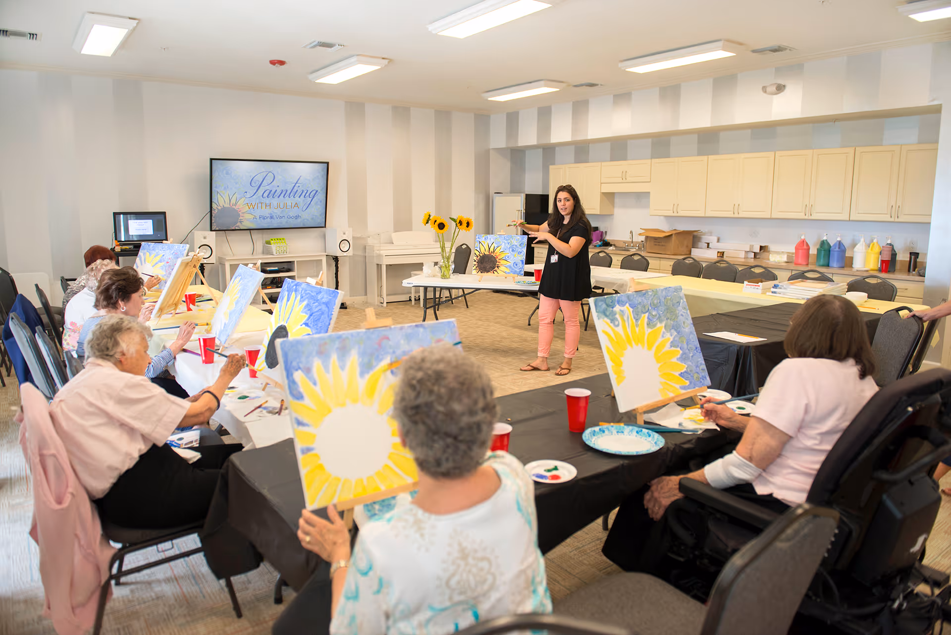 A group of elderly individuals seated around tables in a bright room, participating in a painting class led by a woman standing and instructing them. Each participant has a canvas with a sunflower painting in progress. The room has light-colored walls, cabinets, and art supplies on a counter in the background.