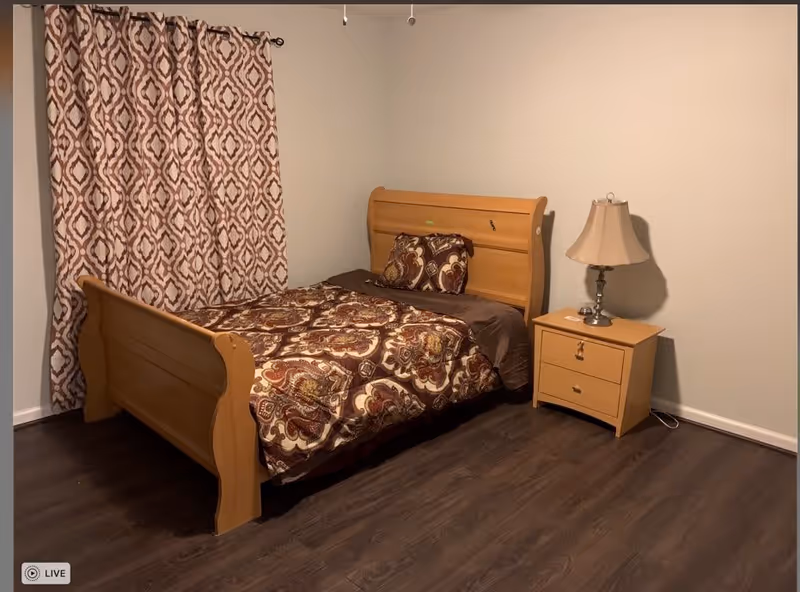 A bedroom in a senior care facility featuring a wooden bed with a patterned brown and beige comforter and matching pillow. Next to the bed is a wooden nightstand with a beige lampshade. The room has dark wood flooring and beige walls with patterned curtains covering the window.