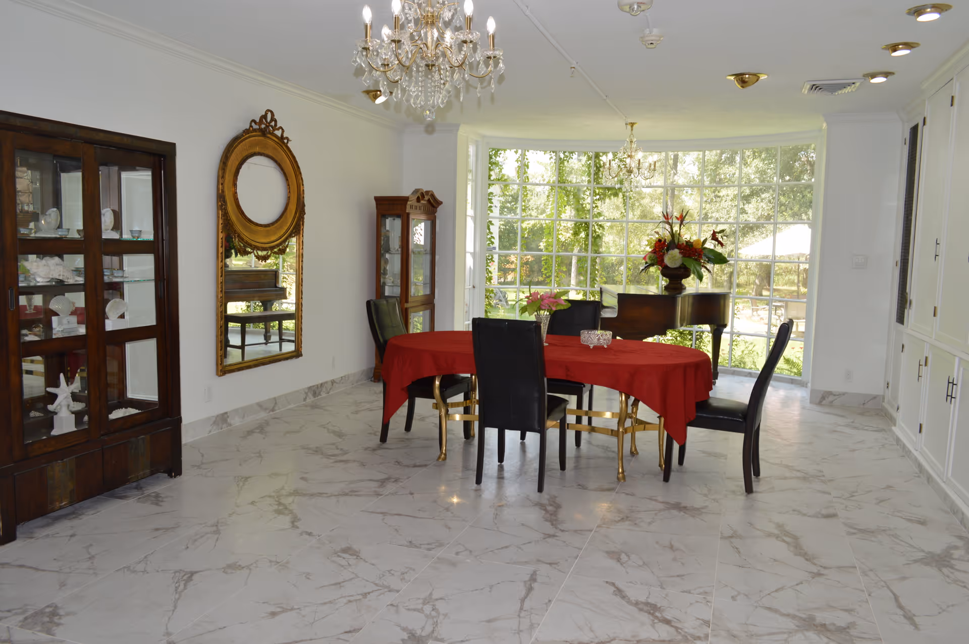 Bright formal dining room with a round table covered in a red tablecloth, black chairs, chandeliers, a grand piano by a large window, and glass-front cabinets.