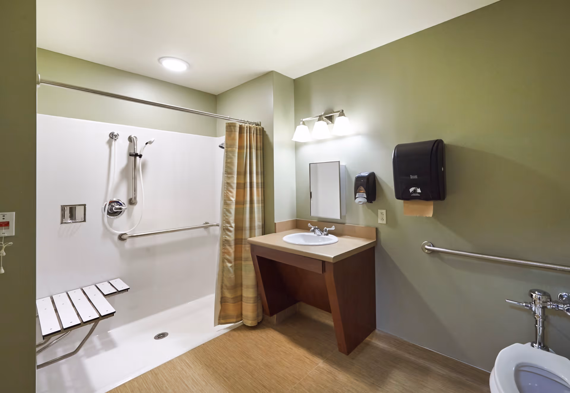 Accessible bathroom with a walk-in shower featuring a fold-down seat and grab bars, a sink with a mirror and light fixture above it, a paper towel dispenser, and a toilet with grab bars on the wall.