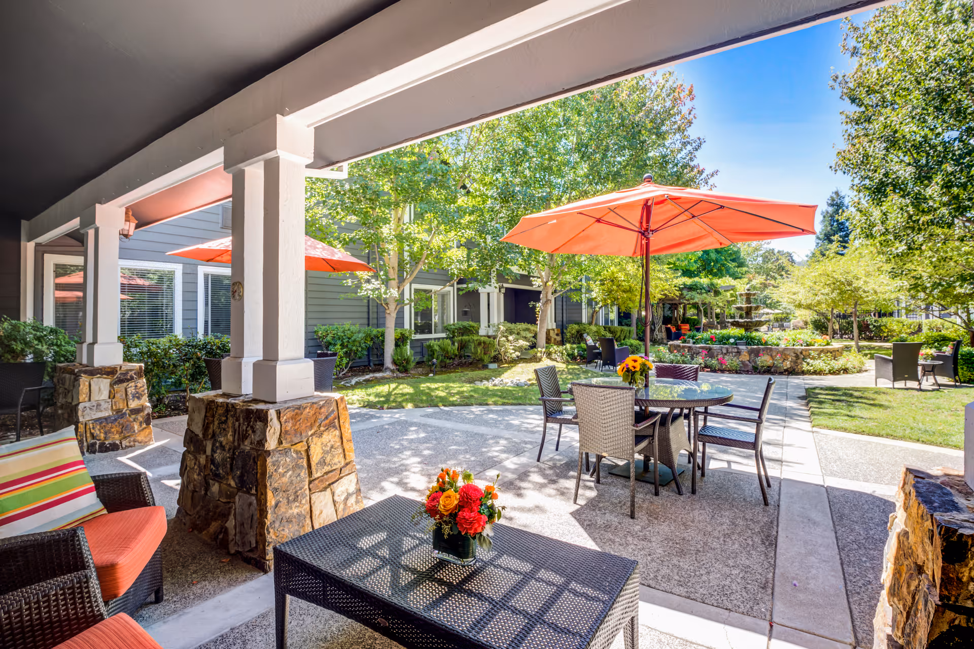 Outdoor patio area at Aegis Living Moraga featuring wicker chairs with orange cushions, a glass-top table with a flower arrangement, and several round tables with orange umbrellas. The patio overlooks a garden with green trees, shrubs, and a stone fountain under a clear blue sky.