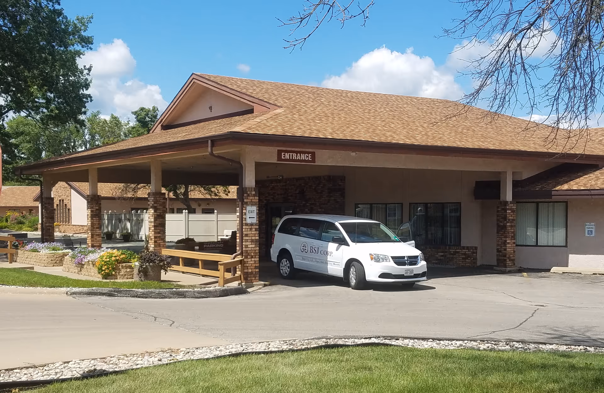 Exterior view of a senior living facility entrance with a covered drop-off area. A white van with the logo 'BSJ Corp.' is parked under the entrance canopy. The building has a brick and beige facade with a brown shingled roof. There are flower beds and green grass in the foreground under a partly cloudy blue sky.