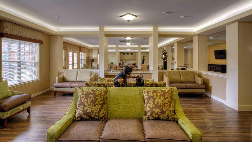 Communal living room with green and brown sofas, patterned pillows, hardwood floors, and decorative sculptures.